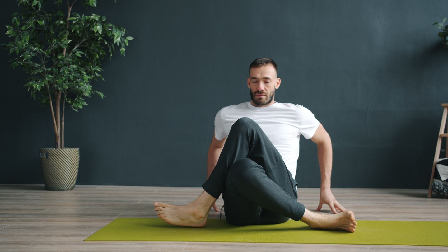 A man in a white shirt doing yoga indoors on a green mat, focusing on relaxation.