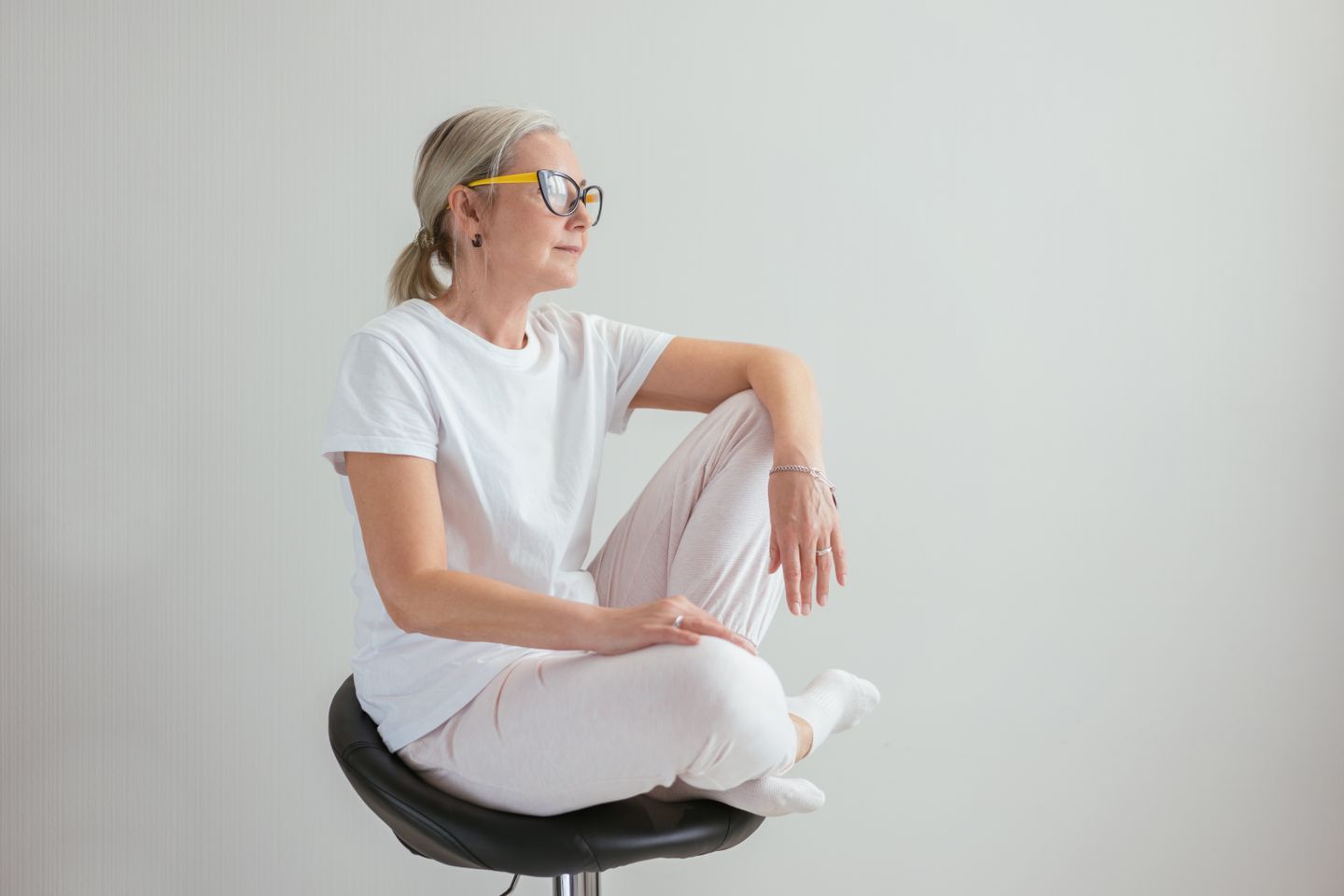 Stylish senior woman in casual wear sitting contemplatively on a modern stool indoors.